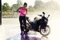 Beautiful stunning girl washes a motorcycle in self service carwash with high pressure water jet in morning at sunrise.