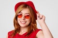 Beautiful stylish female stands sideways, touches fashionable red hat, wears trendy sunglasses, likes red colous, has happy expres