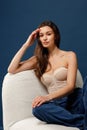 Beautiful stylish girl in blue jeans and beige corset sits on a chair and touches her hair. Blue background Royalty Free Stock Photo