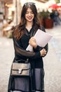 beautiful stylish hipster girl walking in sunny city street, holding magazine. happy gorgeous woman dressed in fashionable outfit Royalty Free Stock Photo