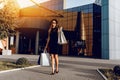 Beautiful stylish Shopaholic girl, in a black dress, black hat and sunglasses, stands with shopping bags in the city against the Royalty Free Stock Photo
