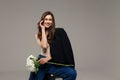 A beautiful stylish smiling girl in blue jeans and a black jacket sits on a chair on a gray background and holds white flowers in Royalty Free Stock Photo