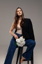 A beautiful stylish smiling girl in blue jeans and a black jacket sits on a chair on a gray background and holds white flowers in Royalty Free Stock Photo