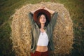 Beautiful woman in hat relaxing on haystack in summer evening field. Tranquility in countryside