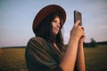 Beautiful stylish woman in hat taking photo of sunset on phone in summer field. Atmospheric moment Royalty Free Stock Photo