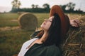 Beautiful stylish woman with herb in mouth relaxing on haystack in summer evening field. Tranquility