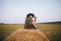 Beautiful stylish woman sitting on haystack and taking photo of sunset on phone in summer field Royalty Free Stock Photo