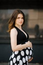 A beautiful stylish young pregnant woman in a black dress with white polka dots holds her hands on her stomach. Happy waiting for Royalty Free Stock Photo