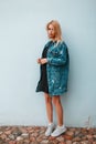 Beautiful stylish young woman blonde in a fashionable long denim jacket in a black trendy dress in sneakers near the vintage wall