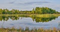 On the smooth water surface of the lake reflected green trees and grass, a blue cloudy sky. Siberia, Russia