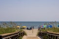 A beautiful summer landscape at the beach with a wooden footpath, people on the beach with colorful umbrellas, blue ocean water