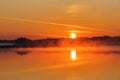 Beautiful sunset is reflecting on the lake. Sky with orange and yellow clouds. Majestic spring morning. Foggy forest