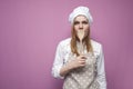 Beautiful surprised girl cook in kitchen clothes with a spoon closes her mouth on a pink background, woman housewife with kitchen