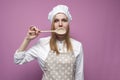 Beautiful surprised girl cook in kitchen clothes with a spoon closes her mouth on a pink background, woman housewife with kitchen