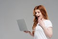Beautiful surprised redhead girl using laptop and gesturing with hand isolated on grey. Royalty Free Stock Photo