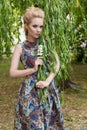 Beautiful sweet tender girl blonde with blue eyes stands near a tree with long branches with a sprig of flowers in her hands Royalty Free Stock Photo