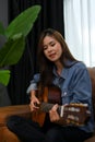 Talented Asian girl in jean jacket singing while playing her acoustic guitar in her living room Royalty Free Stock Photo