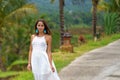 Beautiful tanned woman in white dress posing standing on the road. In the background are palm trees and other tropical vegetation Royalty Free Stock Photo