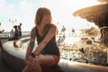 Beautiful tanned young woman with a tattoo on her arm is sitting by the fountain. Sunset in the background. Concept of