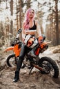 Modern tattooed hipster girl with bright pink hair sitting on her motocross bike in woods