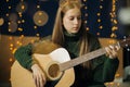 A beautiful teenage girl in Christmas antlers and a green sweater plays guitar