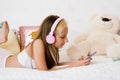 Beautiful teenage girl is relaxing in bed using a touchscreen smartphone, listening to music with a wireless headset, surfing the Royalty Free Stock Photo