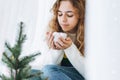 Beautiful teenager girl with cup of cacao with marshmallow sitting on windowsill in the cozy room