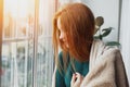 Young woman with long red hair looking through the window at home wrapped in warm comfy blanket Royalty Free Stock Photo