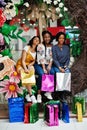 Beautiful three well-dressed afro american girls with colored shopping bags sitting on spring decoration photo zone in mall Royalty Free Stock Photo