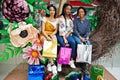 Beautiful three well-dressed afro american girls with colored shopping bags sitting on spring decoration photo zone in mall Royalty Free Stock Photo