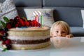 Beautiful three years old toddler boy in blue shirt, celebrating his birthday, blowing candles on homemade baked cake, indoors