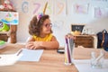 Beautiful toddler wearing glasses and unicorn diadem sitting on desk at kindergarten looking away to side with smile on face, Royalty Free Stock Photo