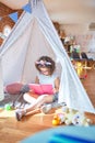 Beautiful toddler wearing glasses and unicorn diadem sitting on the floor inside tipi reading book at kindergarten Royalty Free Stock Photo