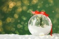 Beautiful transparent Christmas ornament with small fir tree and fairy lights on snow against green background, bokeh effect. Royalty Free Stock Photo