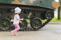 A beautiful two-year-old girl stands on a Sunny day on the street near the huge wheels of a tank, side view. A child next to a tan Royalty Free Stock Photo