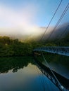 Beautiful view of creek with reflections suspension bridge, blue sky, light clouds, fog, mountains and trees on water Royalty Free Stock Photo