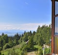 Beautiful view on green forest, blue sky and mountains through a wooden window frame of an old house cottage on a slope of a hill Royalty Free Stock Photo