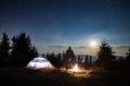 Male hiker looking at bright campfire under night starry sky.