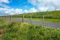 Beautiful view of a small asphalt country road through green meadows with barbed wire fence against blue sky on a sunny day. Royalty Free Stock Photo