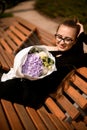 Beautiful view on stylish dressed girl sitting on bench and holding delicate mix of fresh flowers in white craft paper Royalty Free Stock Photo