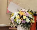 Beautiful wicker basket with lots of white yellow pink flowers with blank white card on blurry background of room. Royalty Free Stock Photo