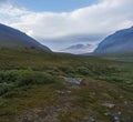 Beautiful wild Lapland nature landscape with blue glacial river, birch tree bushes, snow capped mountains and dramatic clouds.