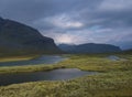 Beautiful wild Lapland nature landscape with blue glacial river, birch tree bushes, snow capped mountains and dramatic clouds.