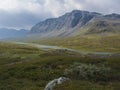 Beautiful wild Lapland nature landscape with blue glacial river, birch tree bushes, snow capped mountains and dramatic clouds.