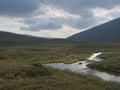 Beautiful wild Lapland nature landscape with blue glacial river, birch tree bushes, snow capped mountains and dramatic clouds.