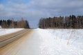 Purified empty suburban sand-strewn road through a snowfield and a forest under blue sky with white clouds on frosty bright sunny