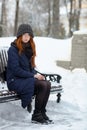 Beautiful Winter Portrait Of Young Adorable Redhead Woman In Cute Knitted Hat Winter Having Fun Sitting On Bench Snowy Park