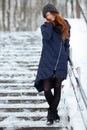 Beautiful winter portrait of young adorable redhead woman in cute knitted hat winter having fun on snowy park stairway