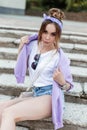 beautiful woman with bandana in trendy casual clothes: white tank top, purple sweatshirt and ripped denim shots sits on the steps