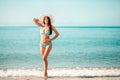 A beautiful woman in a bikini and a beach hat poses standing on the beach. In the background, the sea and sky. Copy space. The Royalty Free Stock Photo